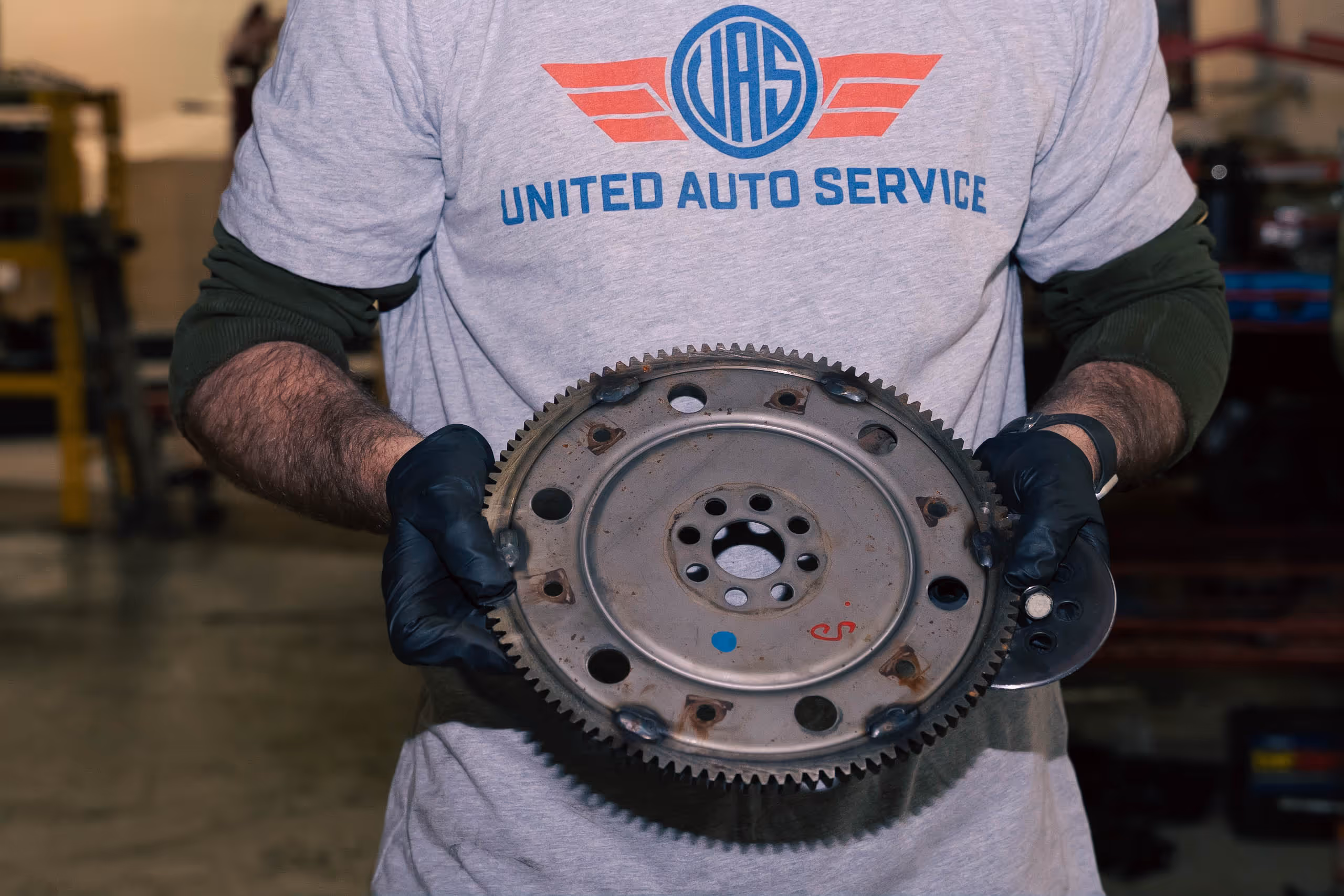 Person wearing a United Auto Service shirt and black gloves holding a metal automotive flywheel gear.