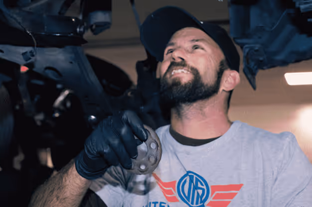 Male mechanic in a cap and gloves inspecting a car part under a vehicle lift in a garage.