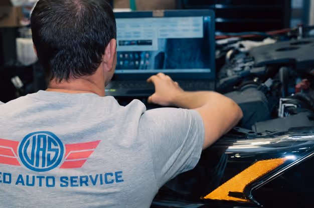 Auto mechanic wearing a gray T-shirt with logo working on a laptop near a car with its hood open in a repair shop.