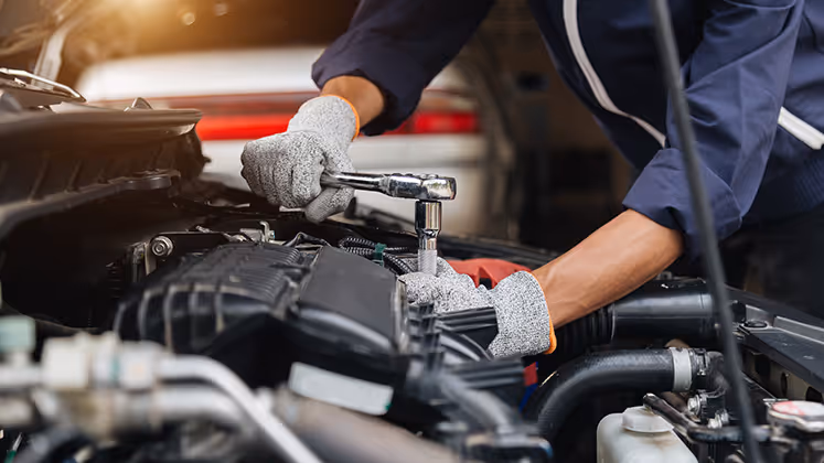 Mechanic wearing gray gloves and a navy jacket using a socket wrench on a car engine under the open hood in a garage.