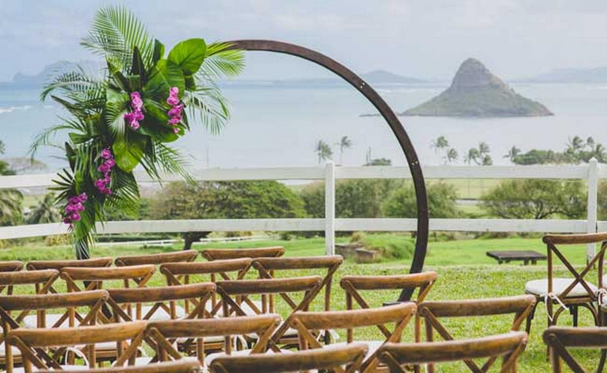 Outdoor wedding ceremony setup at Paliku Gardens featuring wooden chairs and a circular floral arch overlooking Kaneohe Bay and Mokoliʻi, surrounded by tropical palms and lush greenery.