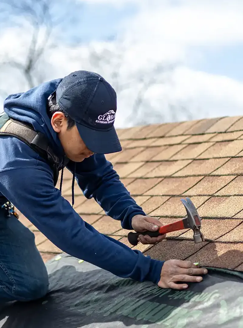 Roofer wearing a cap and hoodie using a hammer to install shingles on a roof.