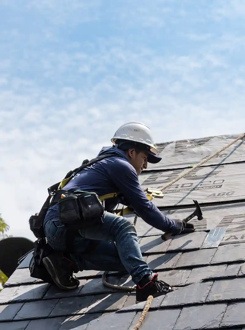Construction worker wearing a safety helmet and harness, hammering roofing nails on a sloped roof.