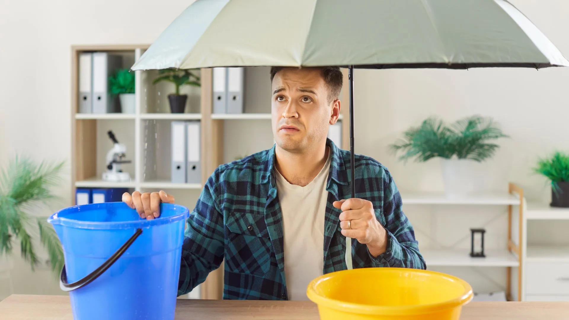 A worried man sitting under a leaking roof with an umbrella and two buckets to catch dripping water