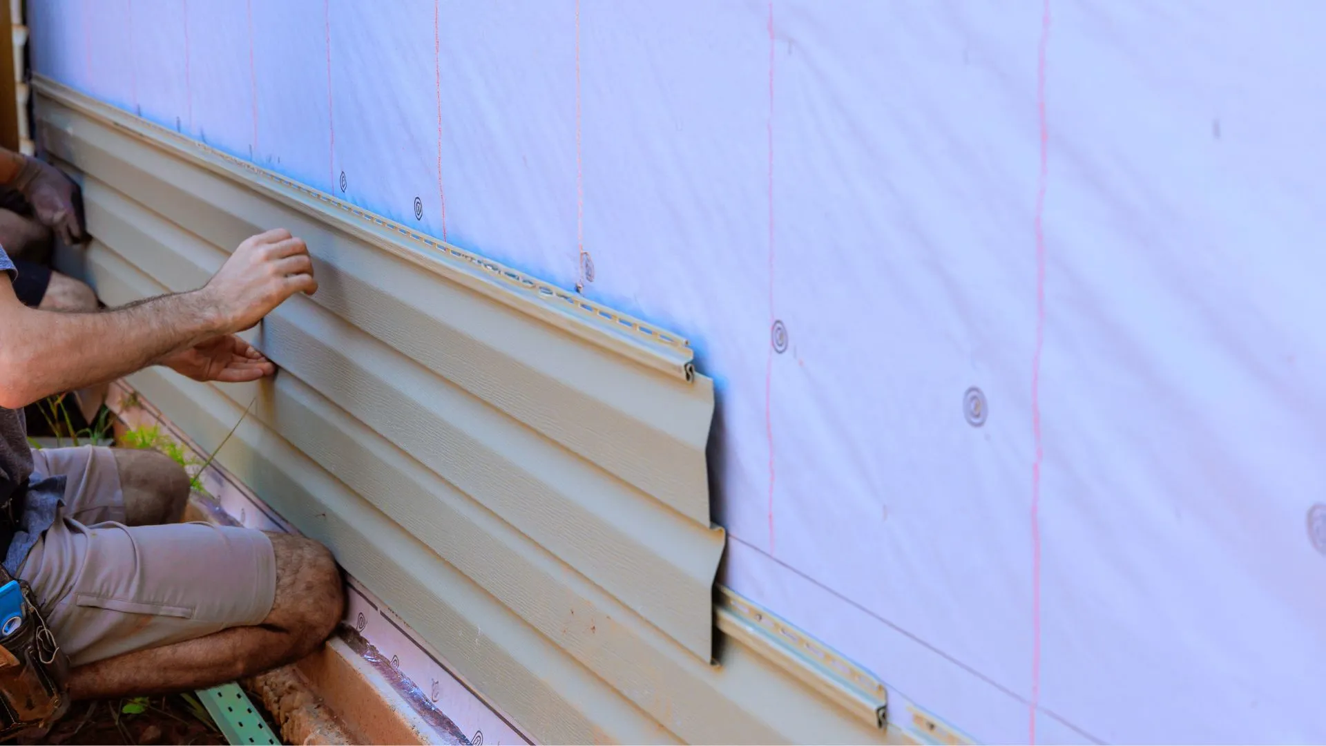 A man wearing shorts working on vinyl siding, with visible white insulation sheets behind the vinyl strips