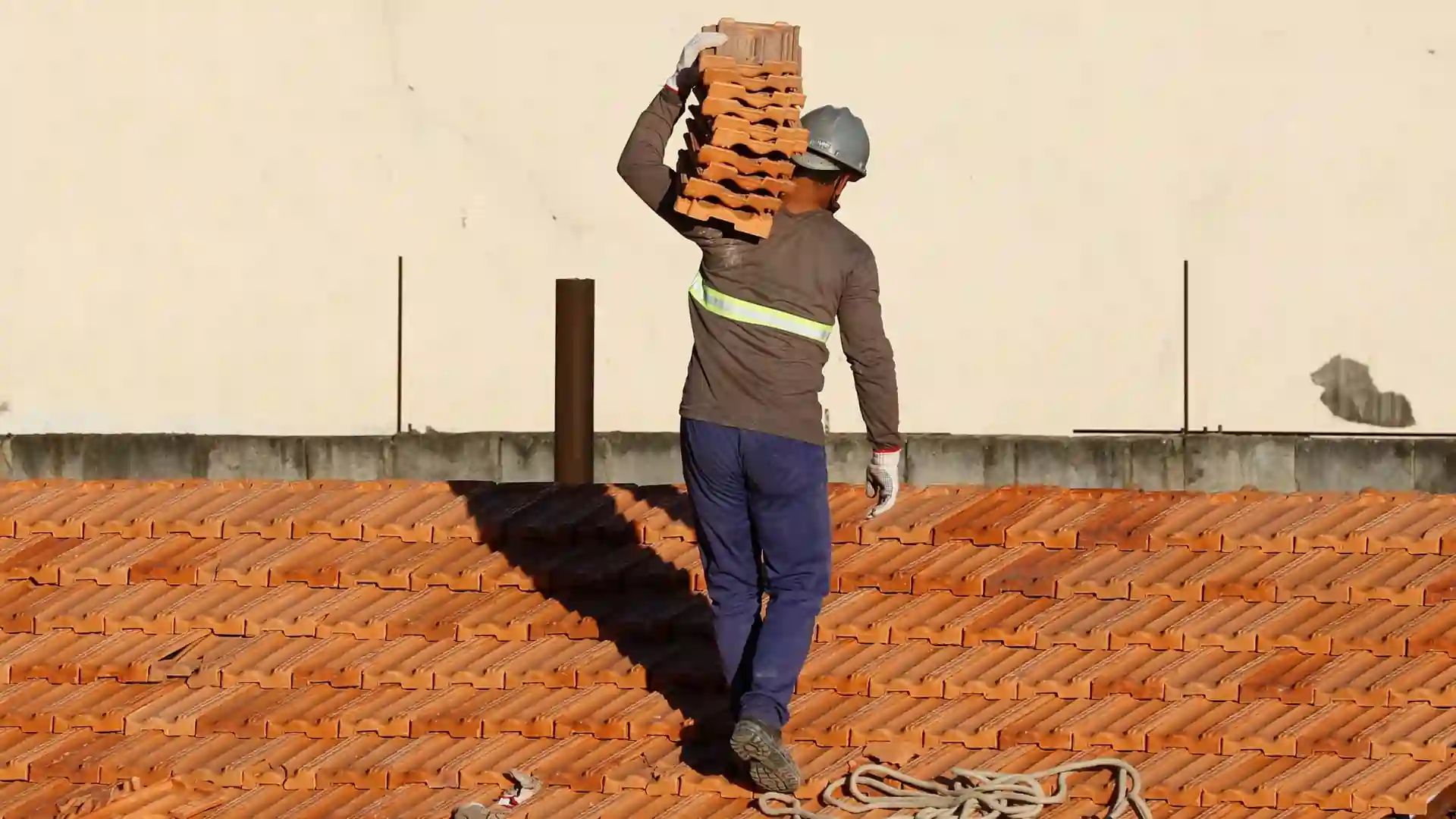 A roofer carrying a heavy tile stack on a roof signifying the labor intensiveness of roofing jobs