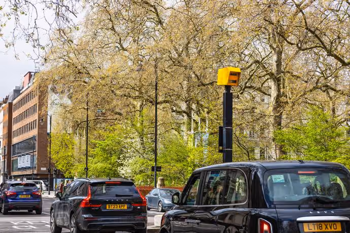 Speed camera in London watching vehicles including a black cab and cars on a leafy street