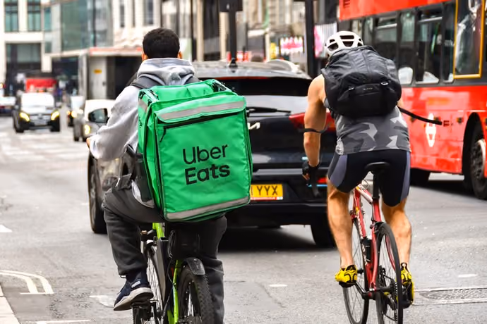 Cyclist carrying an insulated delivery container for the Uber eats cycling alongside a commuer on a street in central London.