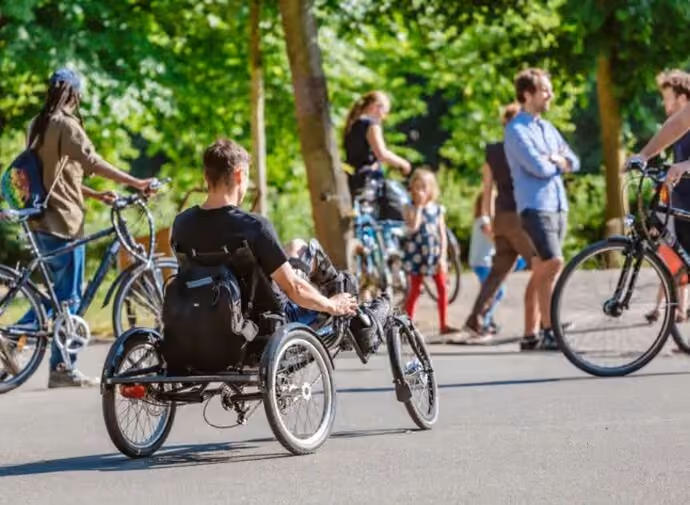 man on trike shares path across park with other cyclists and people on foot. It's summer and the sun is out. 