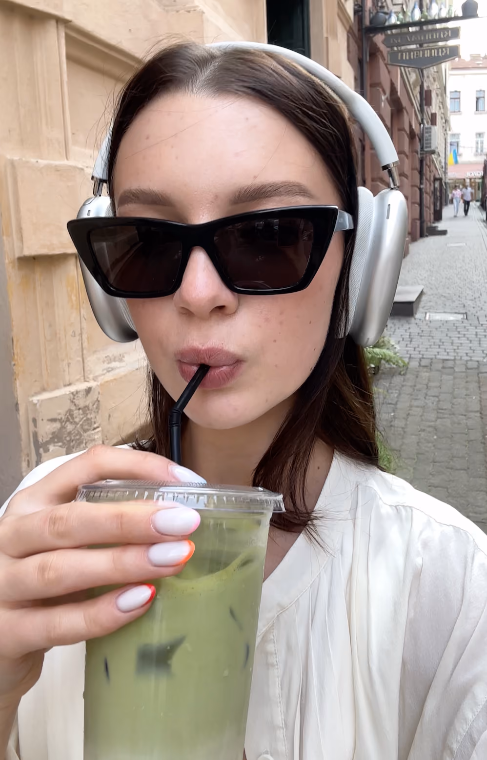 Woman wearing silver headphones and black sunglasses sipping a green iced drink with a black straw on a cobblestone street.