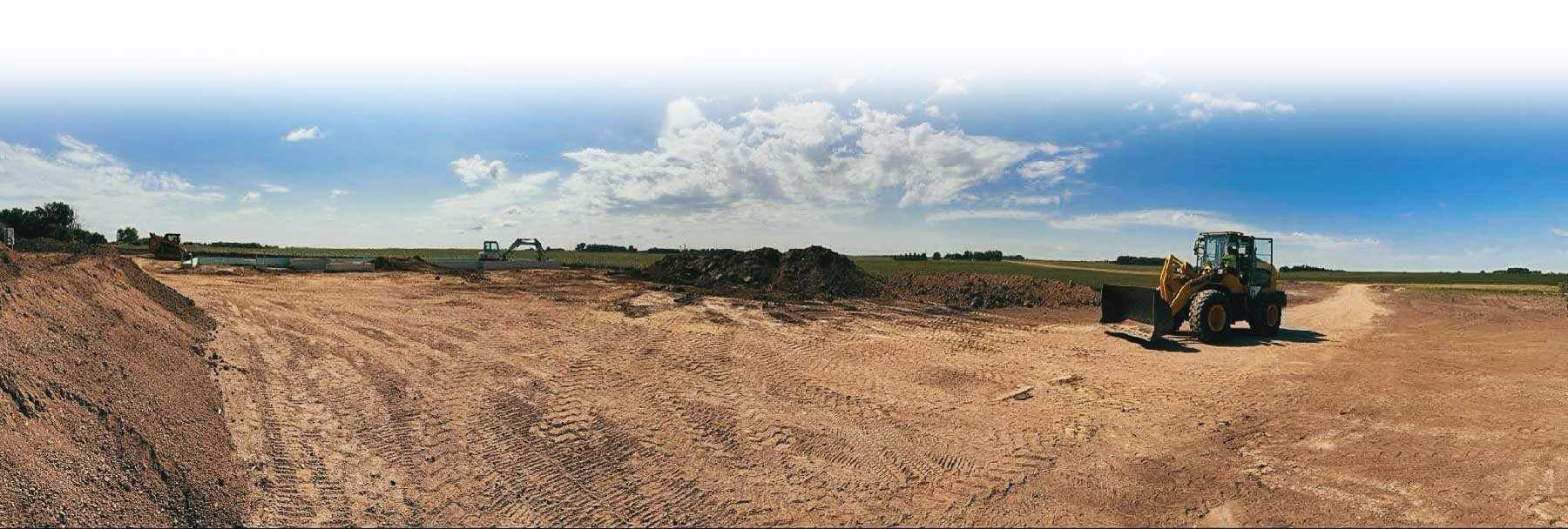 Large open construction site with earthmover and piles of dirt under a partly cloudy sky.