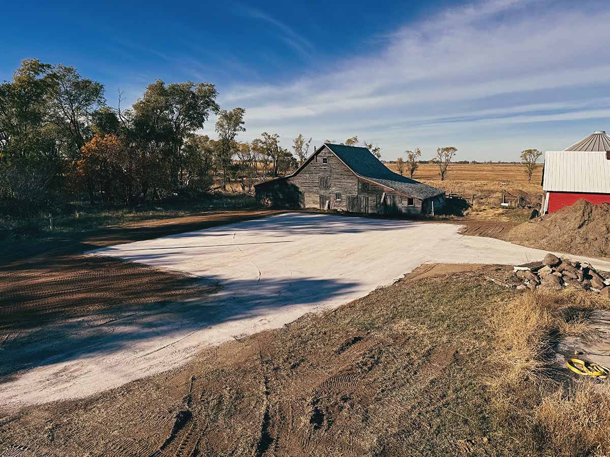 Old wooden barn and a red building with white roof beside a large cleared dirt and gravel area under a blue sky with scattered clouds.