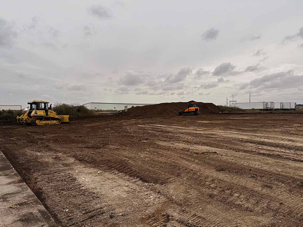 Construction site with a yellow bulldozer on the left and an orange excavator in front of a large dirt mound under a cloudy sky.