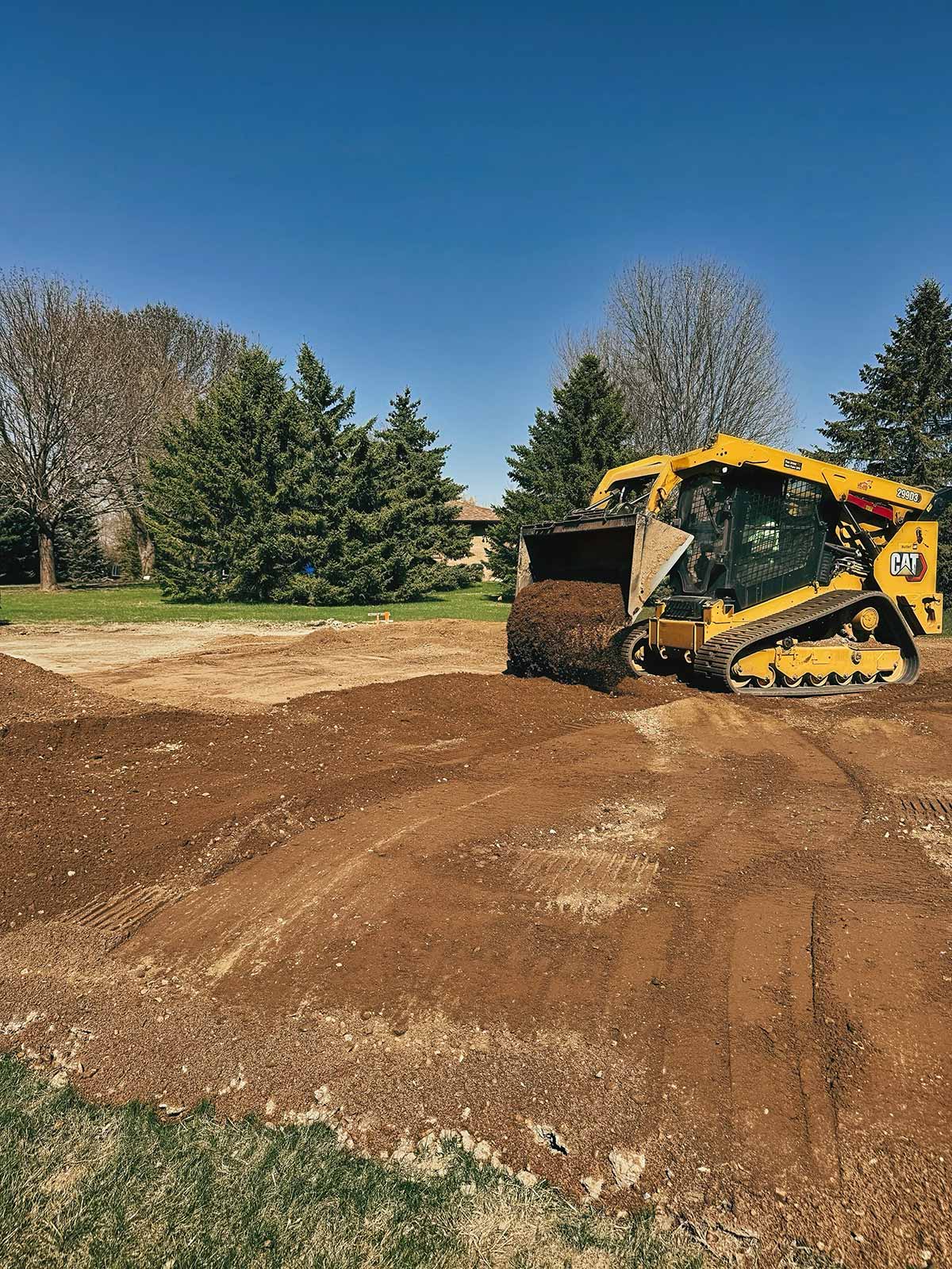 Yellow tracked skid steer loader spreading soil on a cleared area with trees in the background under a clear blue sky.