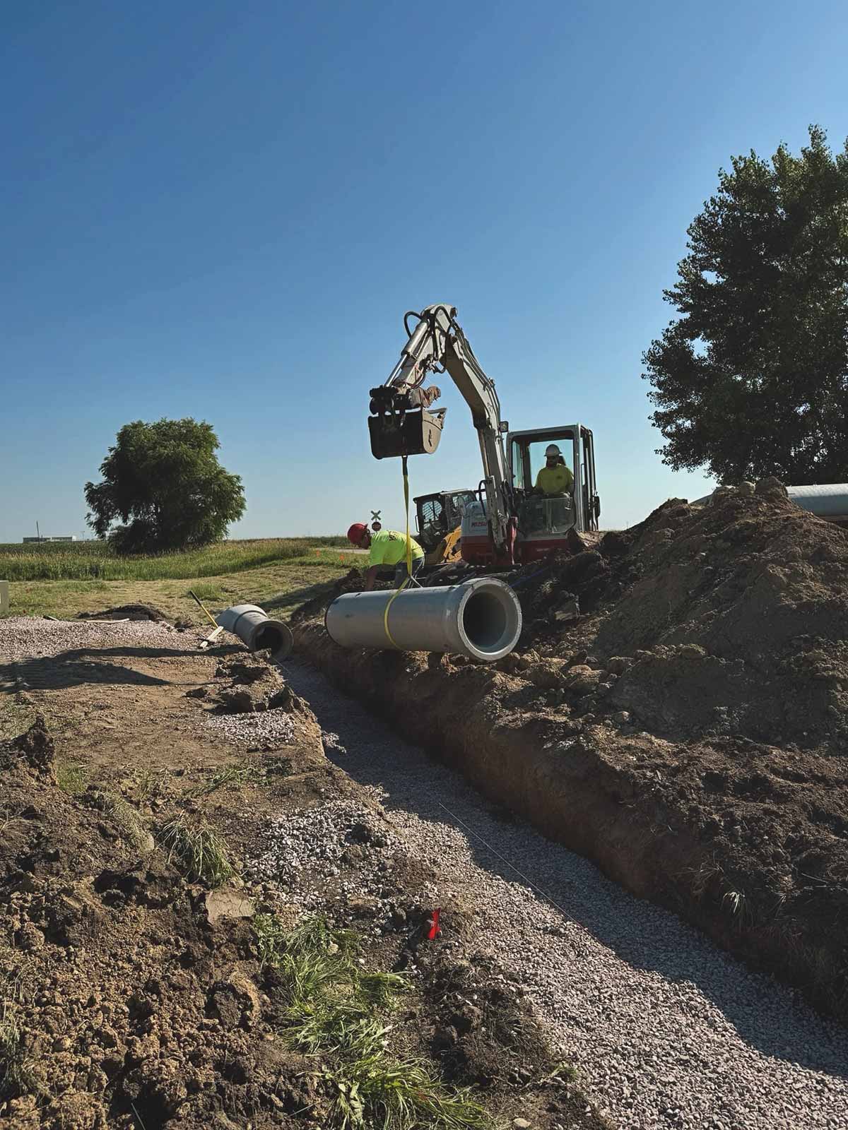 Construction workers using excavator to place a large concrete pipe into a gravel-lined trench under a clear blue sky.