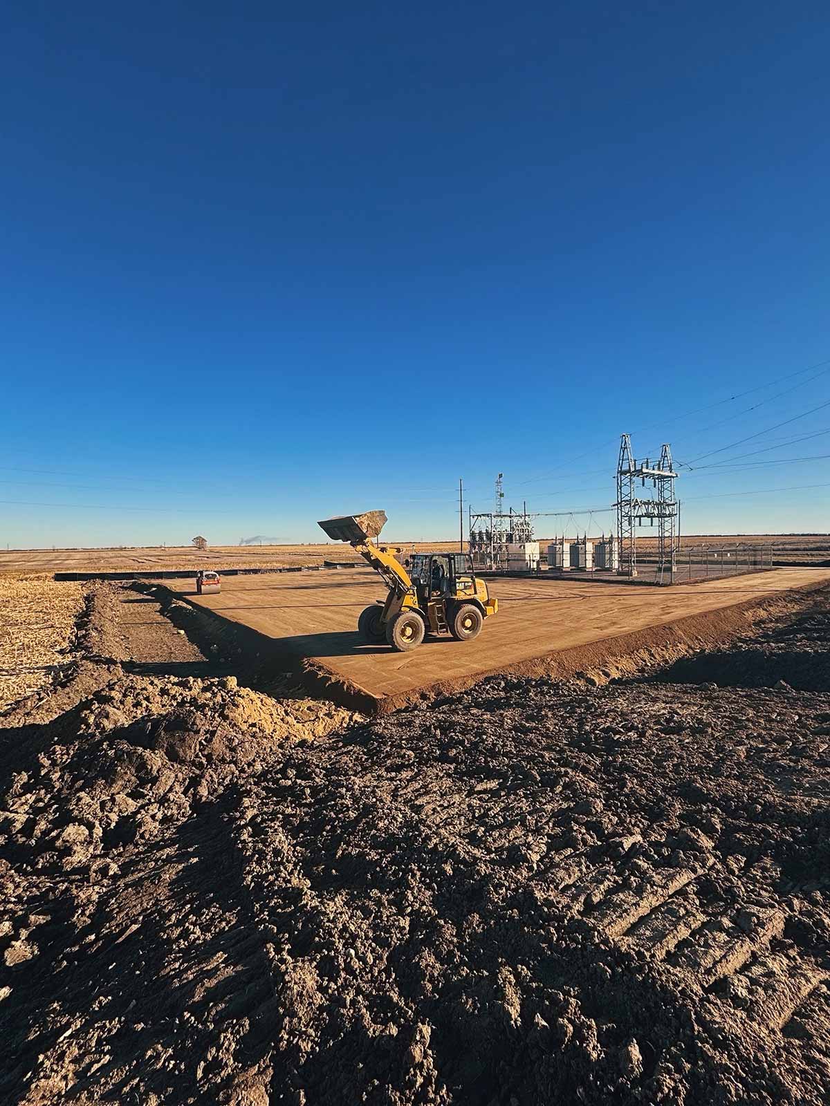 Yellow front loader moving dirt on a construction site near an electrical substation under a clear blue sky.