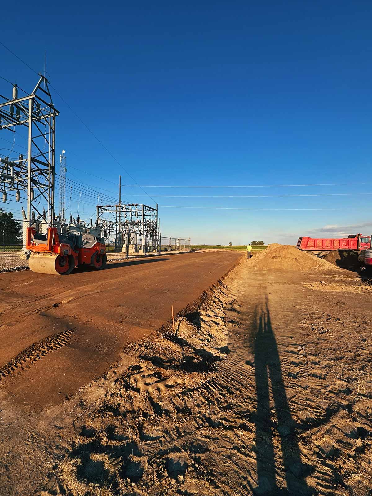 Construction site with dirt road being flattened by steamroller near electrical substation and long shadows cast by a person and equipment.