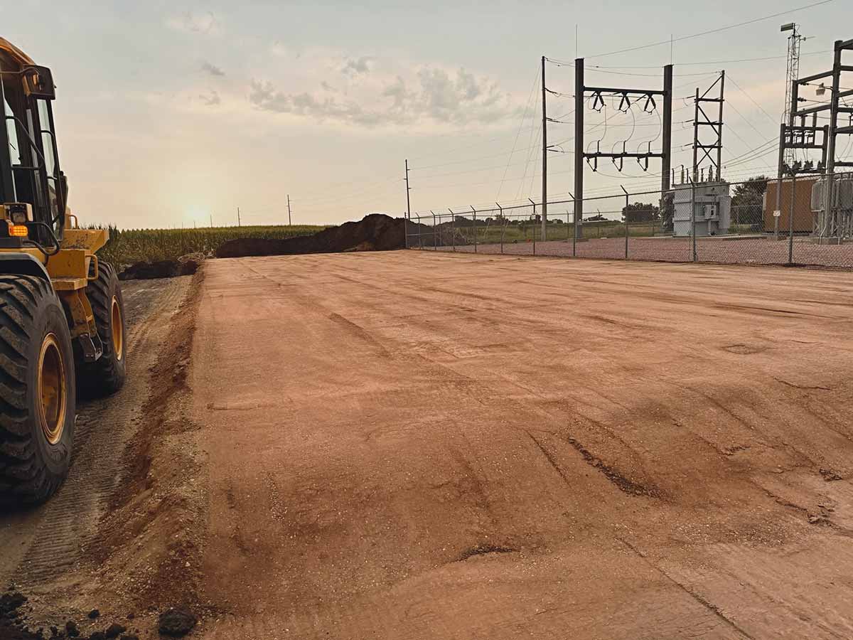Construction site with freshly leveled dirt, a yellow bulldozer on the left, and electrical substation fenced on the right.