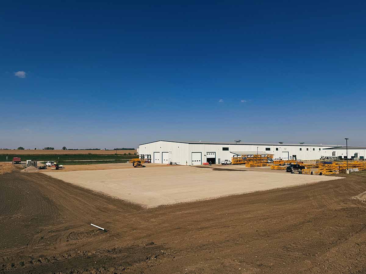 Large white industrial warehouse with yellow construction equipment and vehicles on a dirt and gravel lot under a clear blue sky.