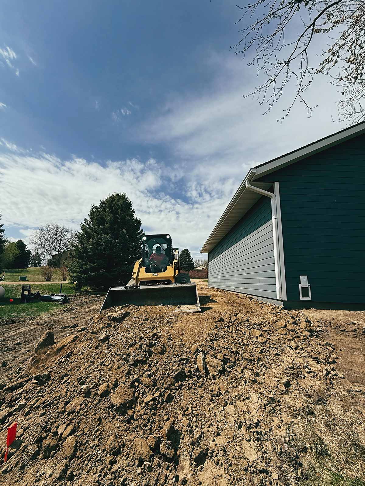 Yellow bulldozer moving dirt beside a blue house under a partly cloudy sky.