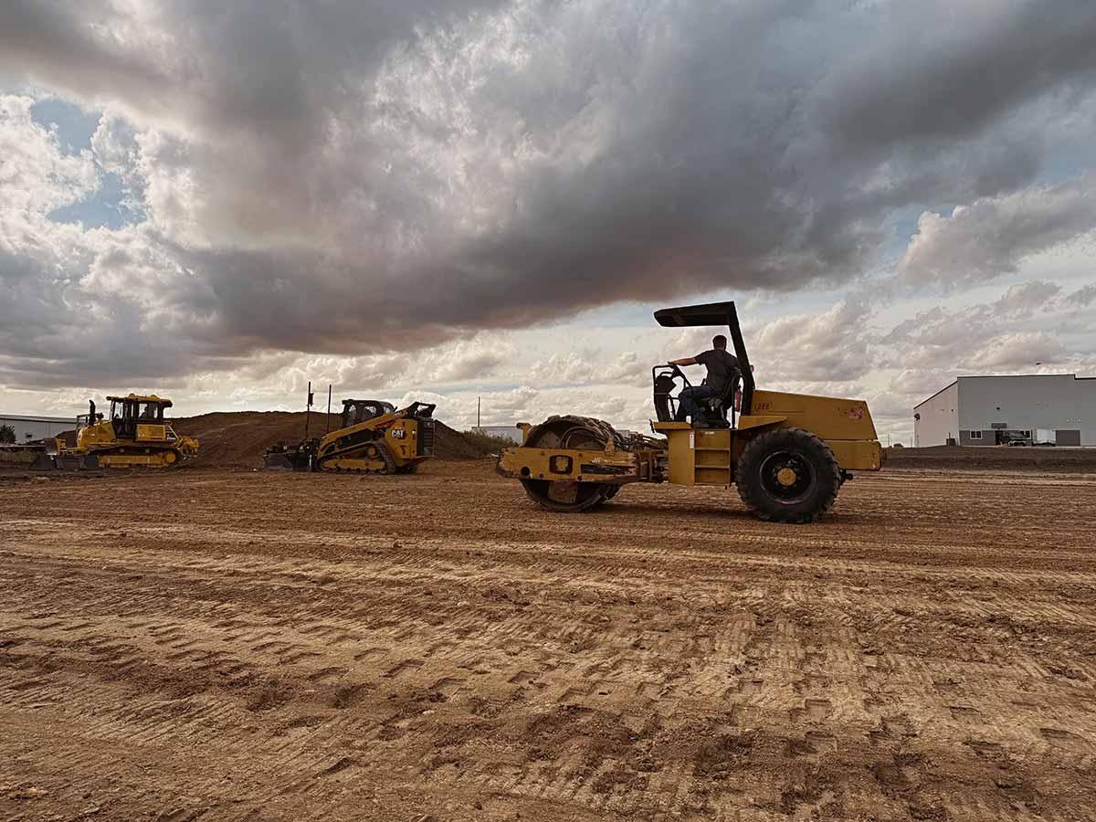 Construction site with heavy machinery including a roller compactor, bulldozer, and skid steer loader under a cloudy sky.