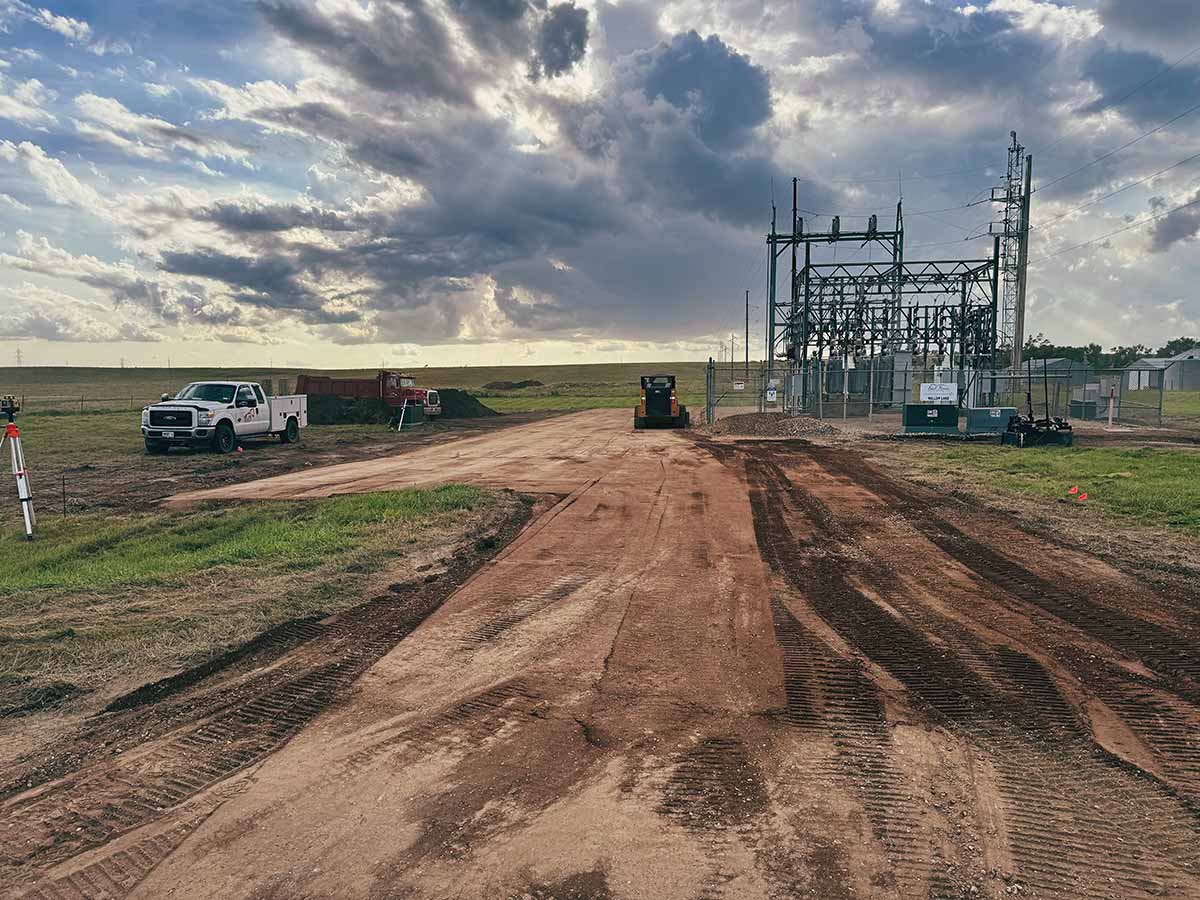 Construction site with dirt road showing tire tracks, a white utility truck, a red dump truck, a bulldozer, and electrical substation under a dramatic cloudy sky.