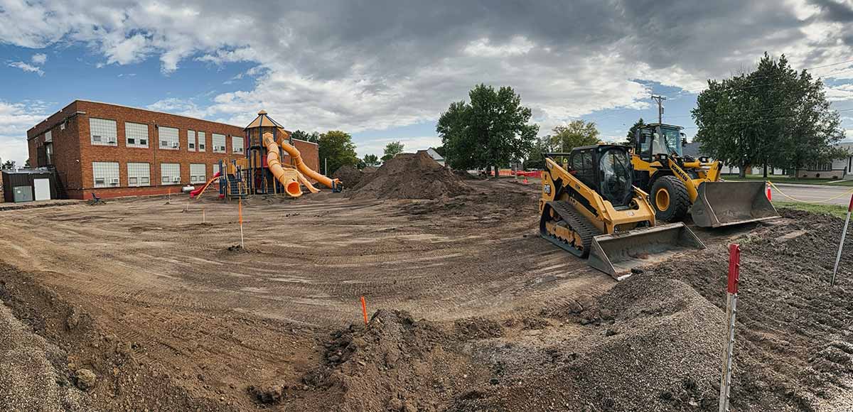 Construction site with earth-moving equipment near a playground and a red brick school building under a cloudy sky.