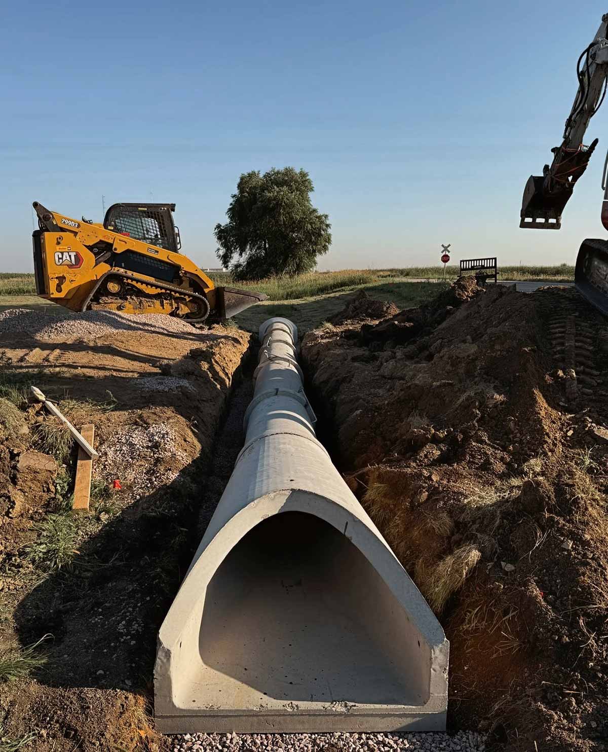 Concrete drainage pipe being installed in a trench at a construction site with heavy machinery and a tree in the background.