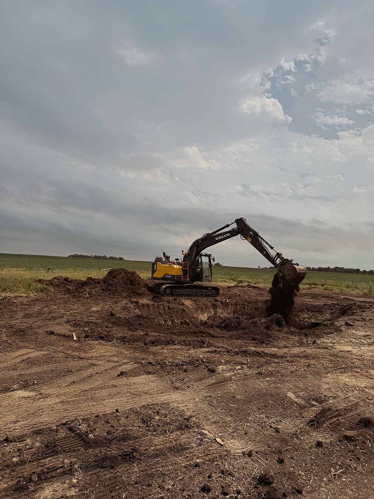 Yellow and black Volvo excavator digging soil in an open field under a cloudy sky.