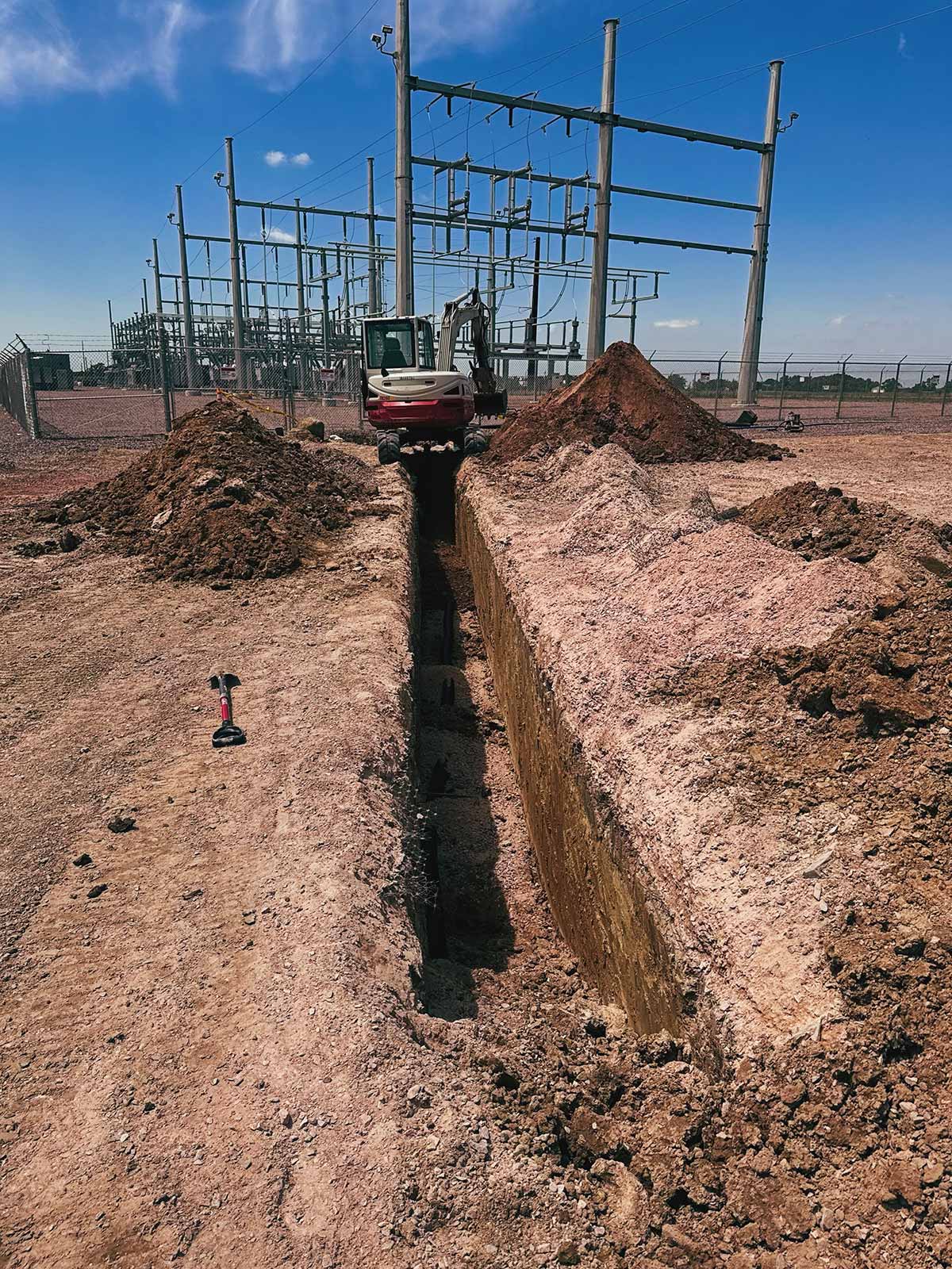 Excavator digging a long trench at an electrical substation with piles of dirt on each side under a clear blue sky.