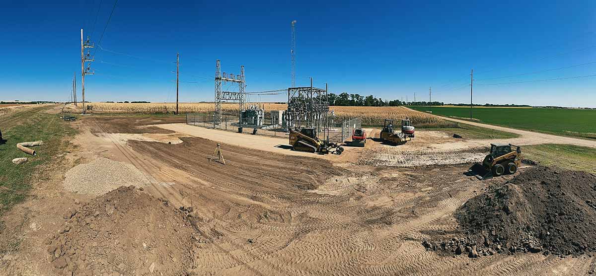 Rural construction site with heavy machinery and power substation equipment under a clear blue sky, surrounded by fields.