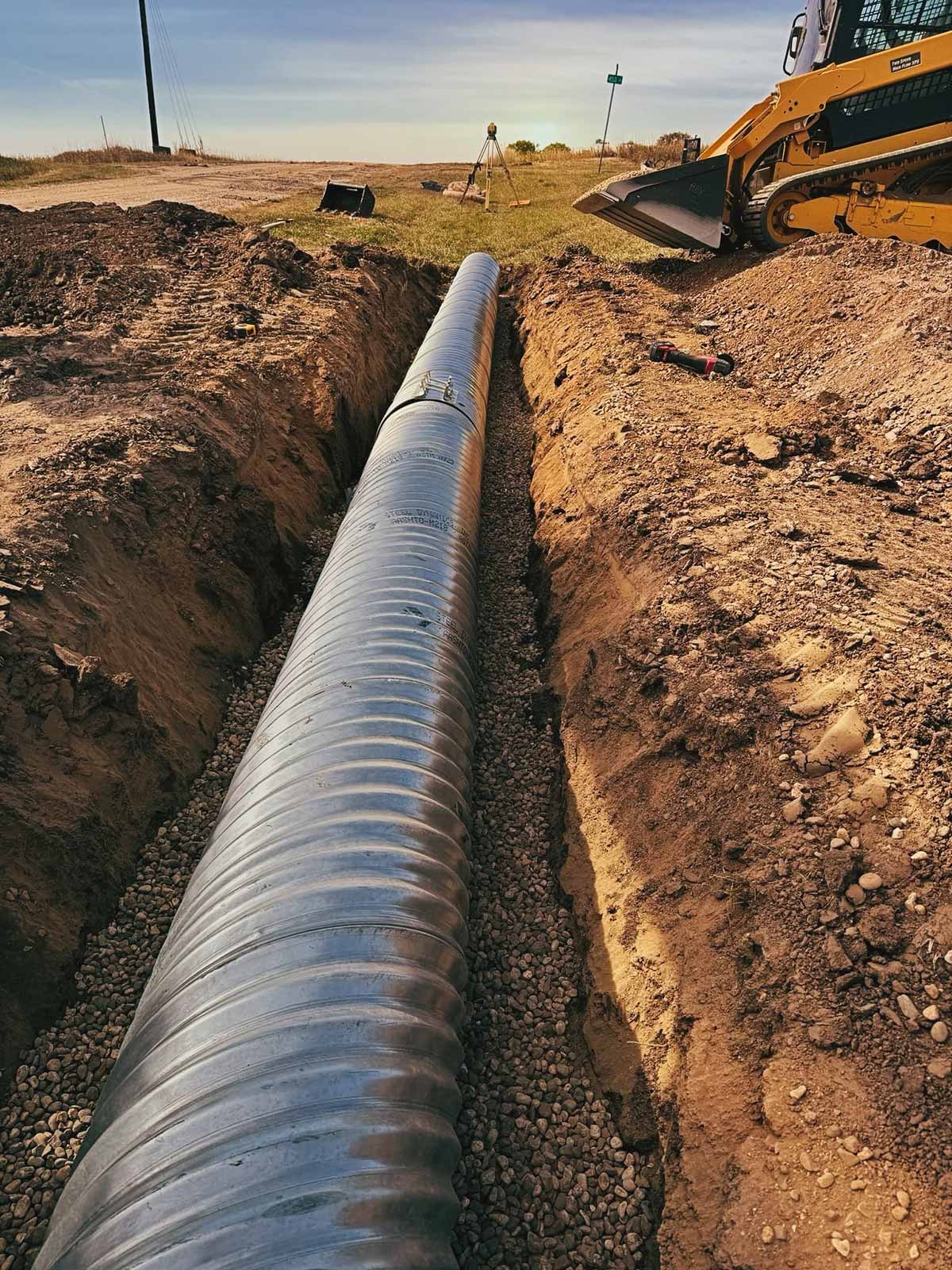 Large corrugated metal drainage pipe laid in a trench with gravel base at a construction site with a skid steer loader nearby.