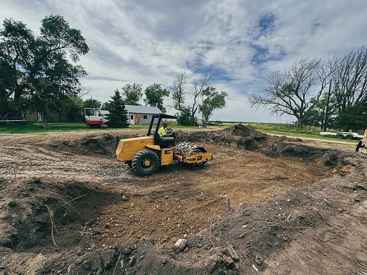 Yellow soil compactor operating inside a large dirt excavation site surrounded by trees and a small building in the background.
