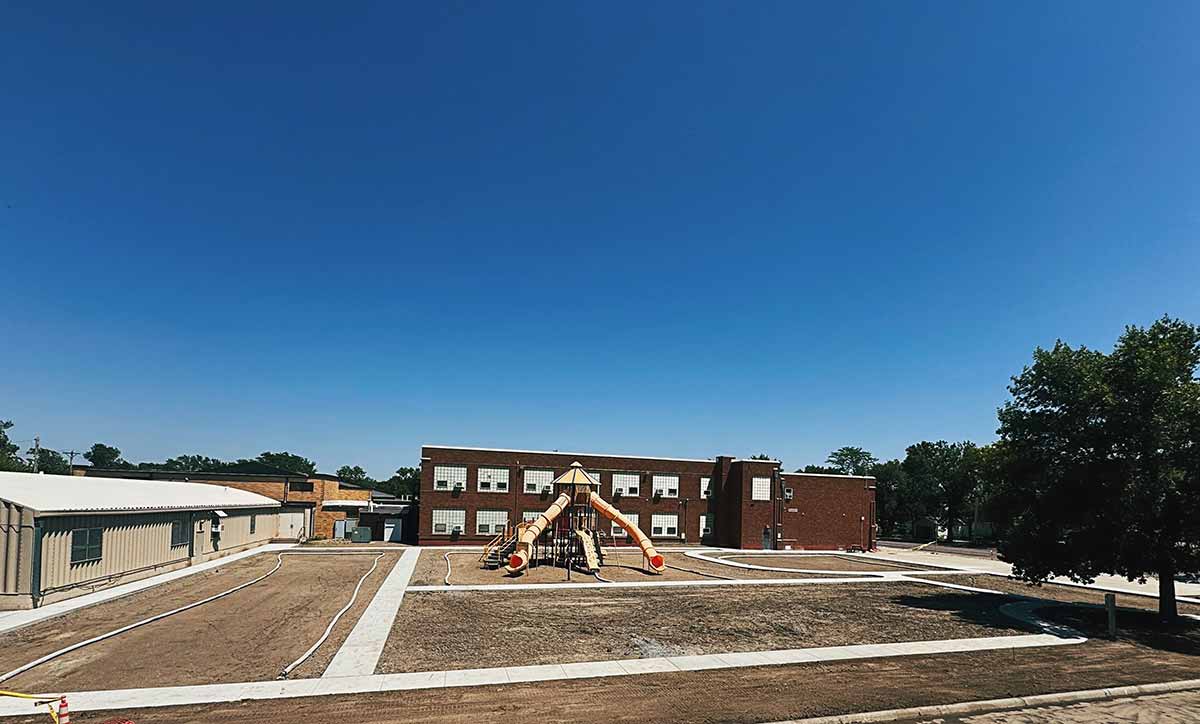 Empty school playground with slides in front of a two-story brick school building under clear blue sky.