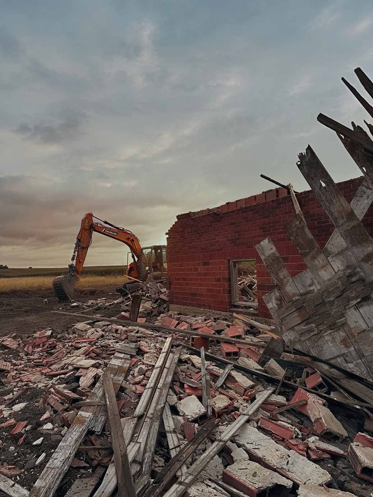 Excavator demolishing a partially torn-down brick building with rubble and wooden debris scattered in the foreground under a cloudy sky.