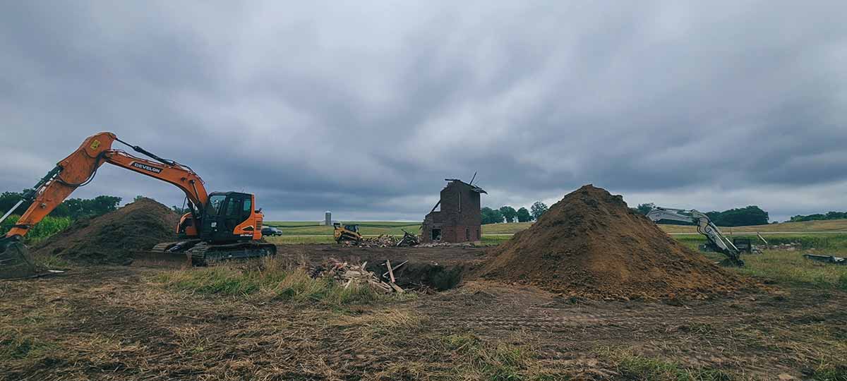 Construction site with excavators and large dirt piles around a partially demolished brick building under a cloudy sky.