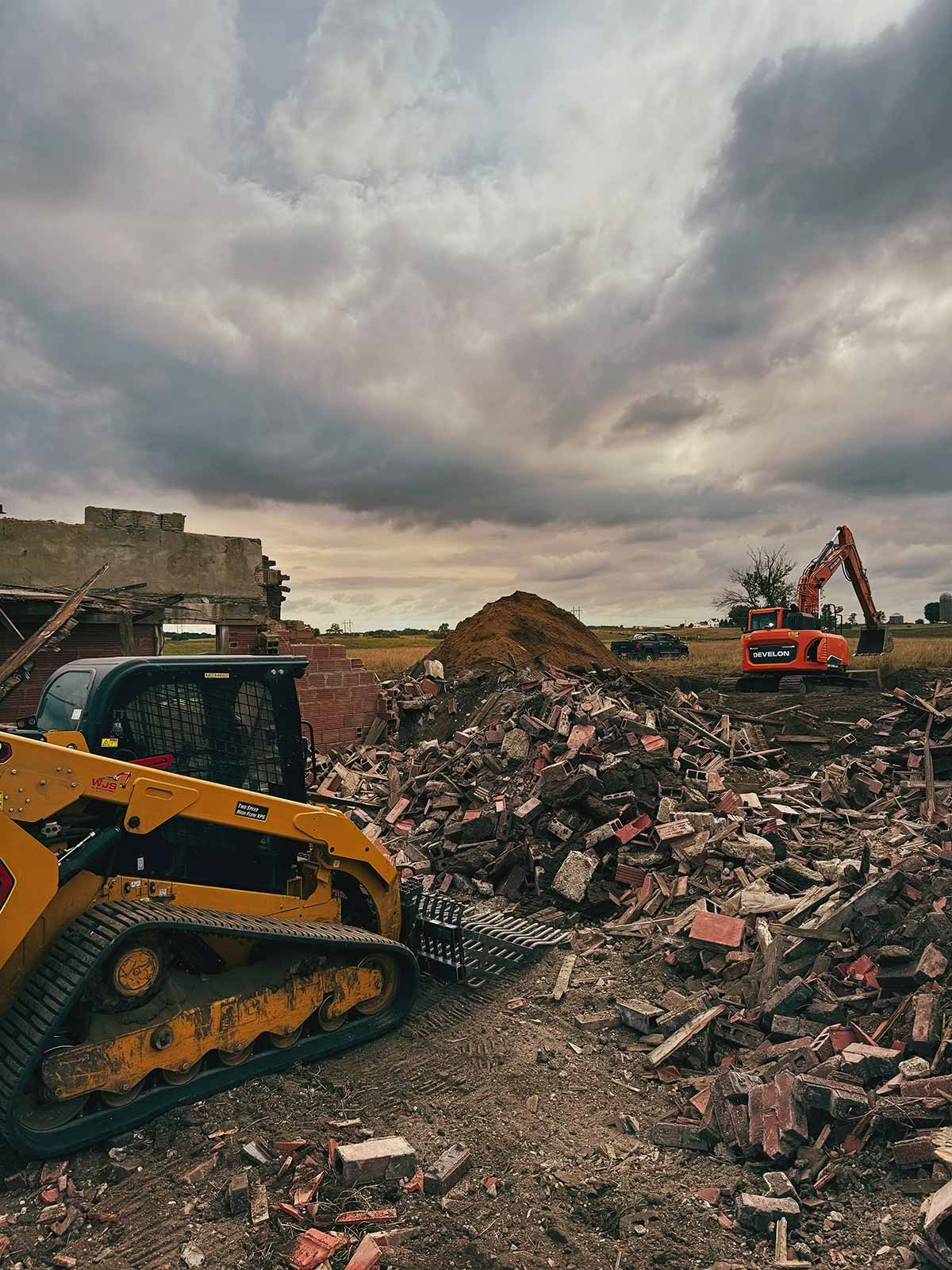 Construction site with heavy machinery among piles of broken bricks and rubble under a cloudy sky.