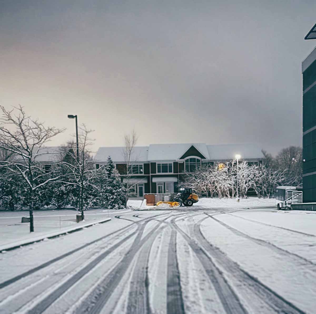 Snow-covered parking lot with fresh tire tracks and a snowplow in front of a building during dusk.