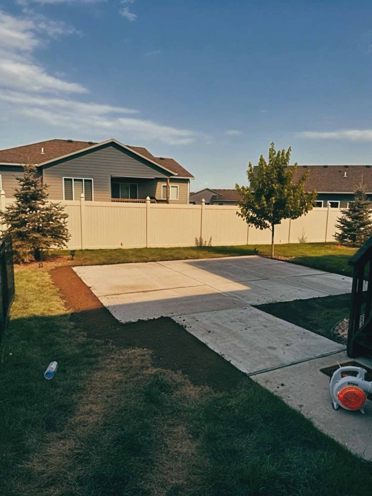 Backyard with a concrete patio and sidewalk, surrounded by a beige fence, grass, two pine trees, one deciduous tree, and a leaf blower on the right side.