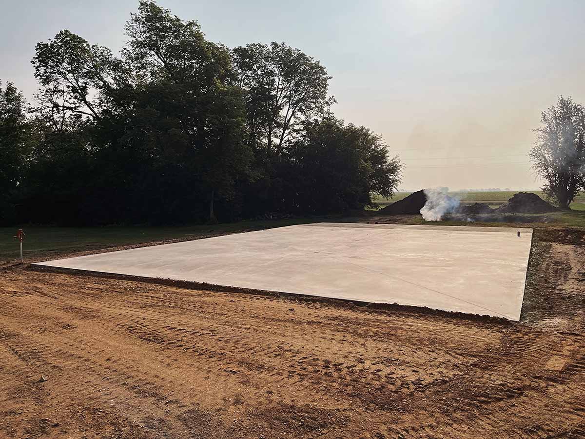 Freshly poured concrete slab foundation on dirt ground with trees and smoke in the background.