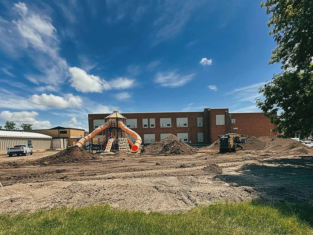 Playground with slides under construction in front of a brick school building on a partly cloudy day.