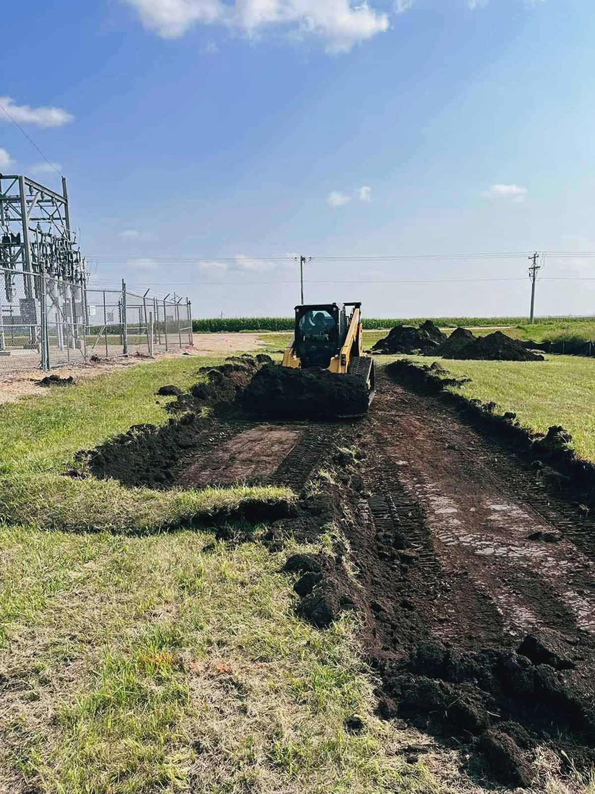 Yellow bulldozer clearing a dirt path in a grassy field with mounds of soil on the sides and power lines in the background.