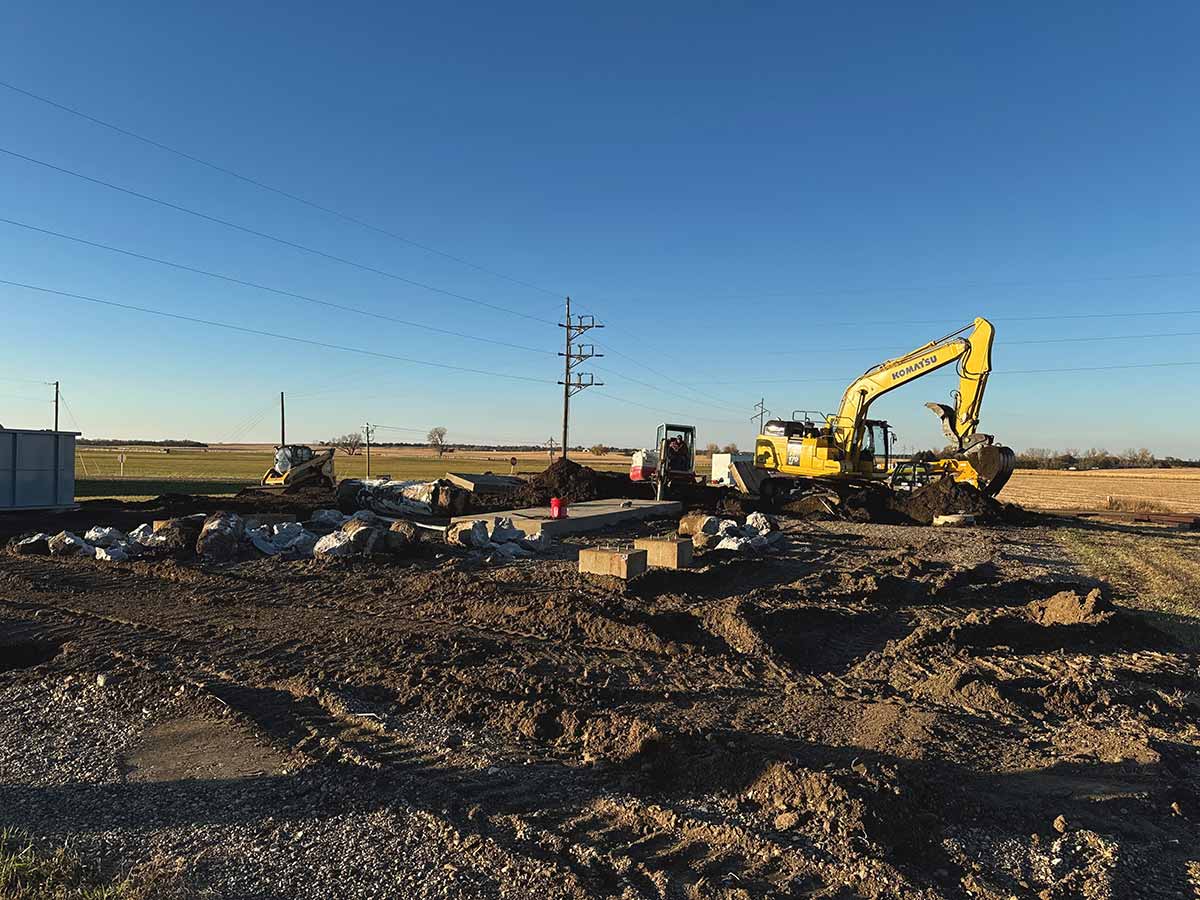 Construction site with a yellow Komatsu excavator digging dirt and piles of wrapped materials on a clear day in a rural area.
