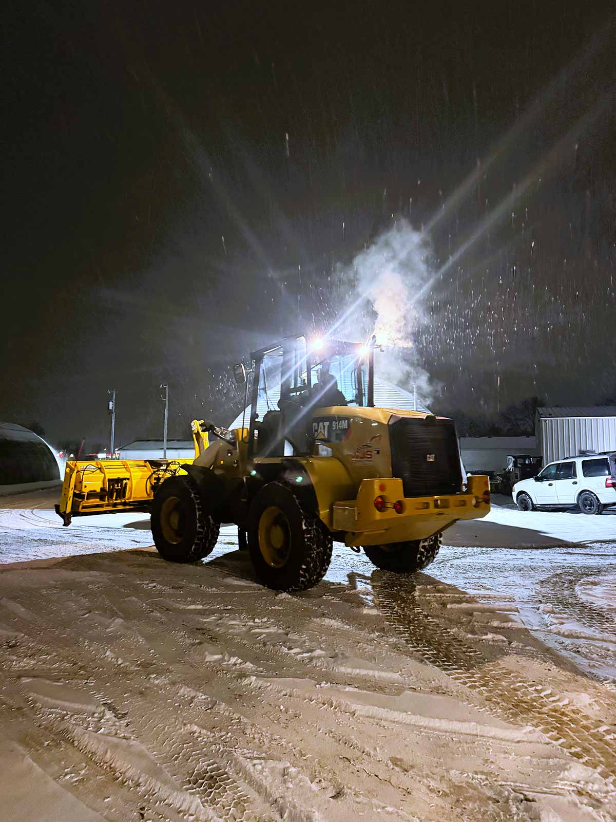Yellow Caterpillar 914M snowplow clearing snow at night with headlights on and snow falling.