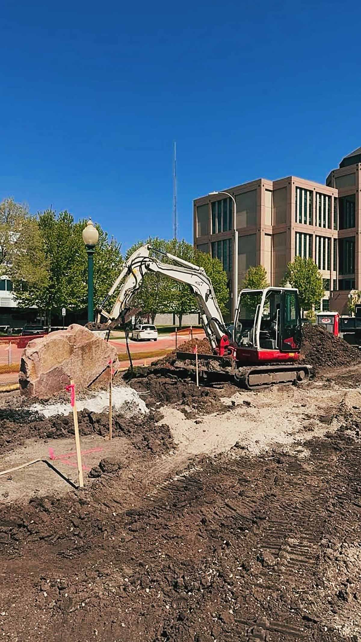 Small excavator moving a large rock at an urban construction site with a multi-story office building and trees in the background.
