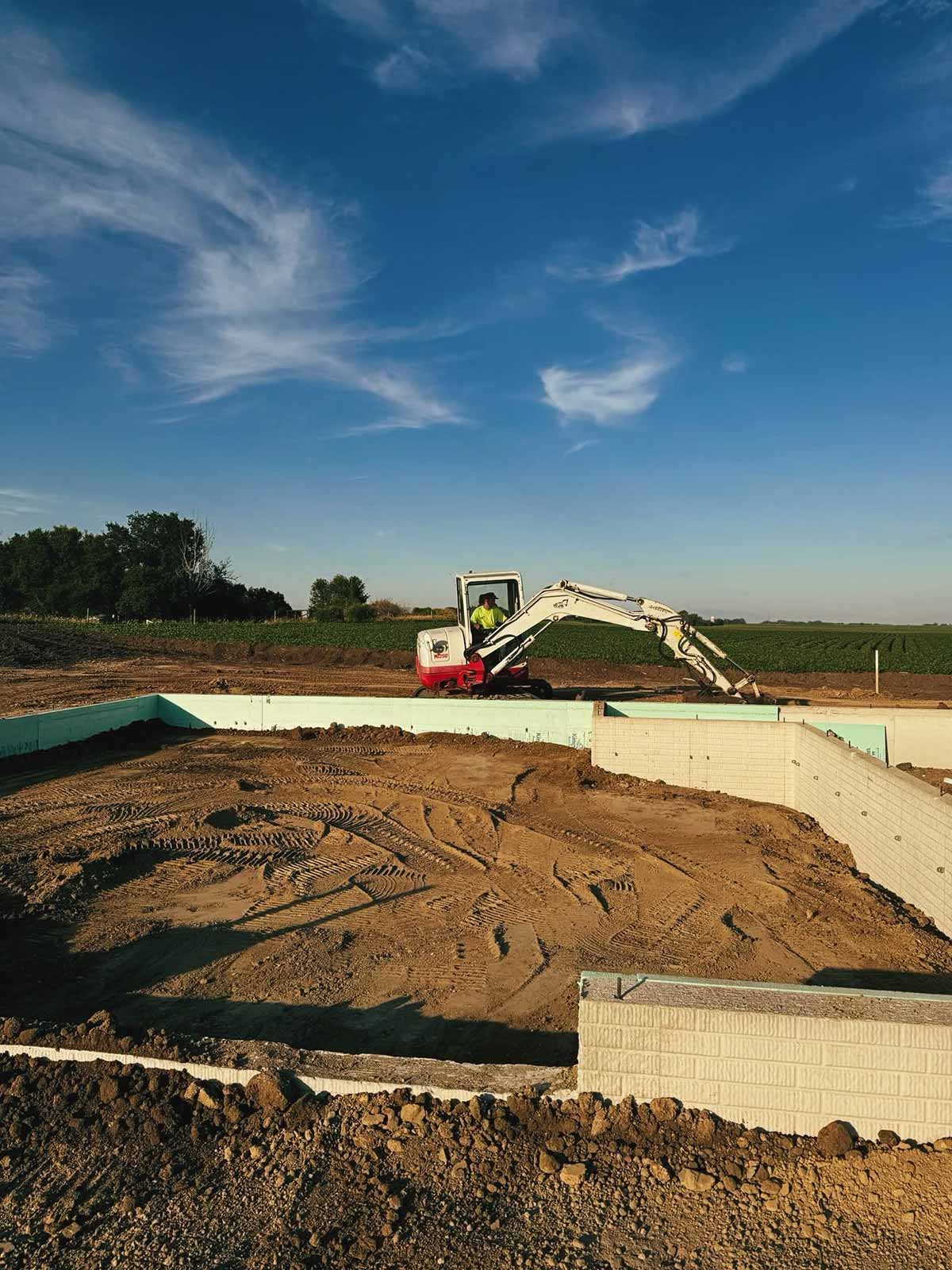 Construction site with excavator digging inside a foundation under a blue sky with wispy clouds.