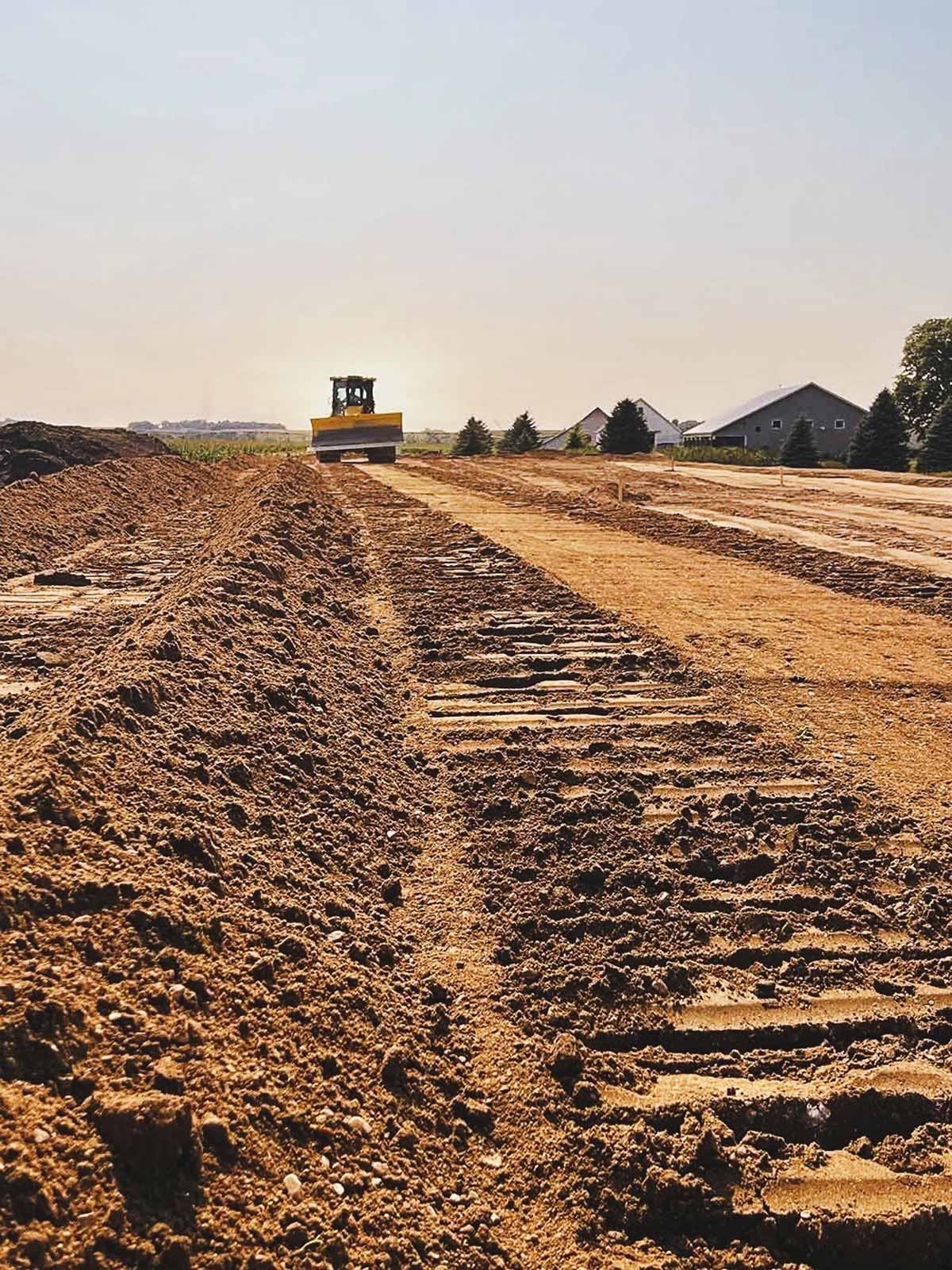 Bulldozer grading a large dirt field with houses and trees in the background under a clear sky.