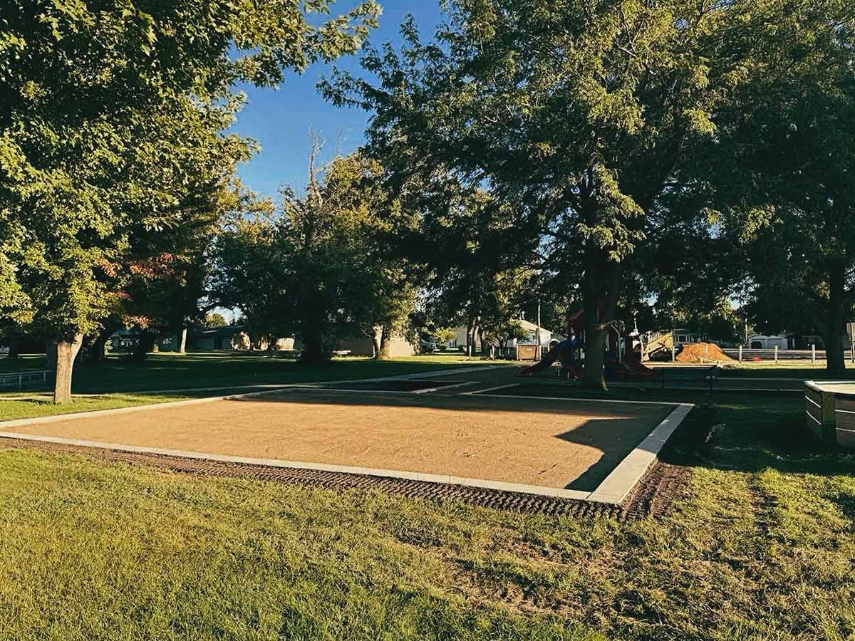 Empty rectangular sandpit in a grassy park area surrounded by large trees and playground equipment.