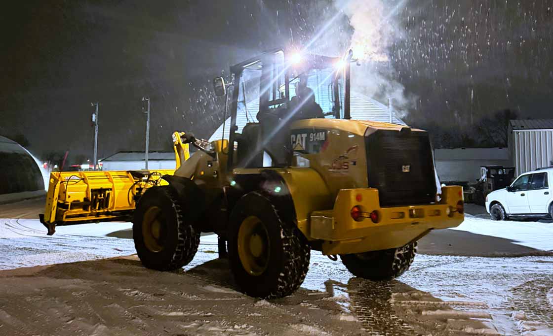 Yellow CAT 914M snowplow clearing snow at night with bright headlights illuminating falling snow.