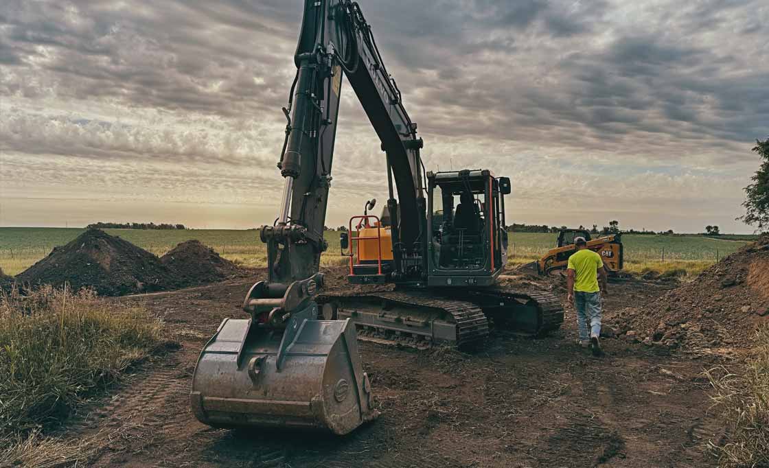 Construction site with a large excavator and a worker in a yellow shirt walking nearby on dirt ground under a cloudy sky.